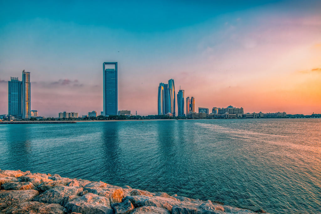 Skyline von Abu Dhabi bei Sonnenuntergang,Abendstimmung,Farbenspiel | Das Bild zeigt einen Blick auf die Skyline von Abu Dhabi (Vereinigte Arabische Emirate) vom Ufer aus, im Vordergrund sind die Wellenbrechersteine am Wasser zu sehen.Links dominiert das markante, hohe Gebäude der ADNOC Headquarters (Abu Dhabi National Oil Company) mit seiner charakteristischen Öffnung an der Spitze, daneben reihen sich weitere moderne Wolkenkratzer ein.Die Aufnahme fängt die Stadt im warmen Licht des Sonnenuntergangs oder der Dämmerung mit einem blauen und orangefarbenen Himmel ein - Realisiert mit Pictrs.com