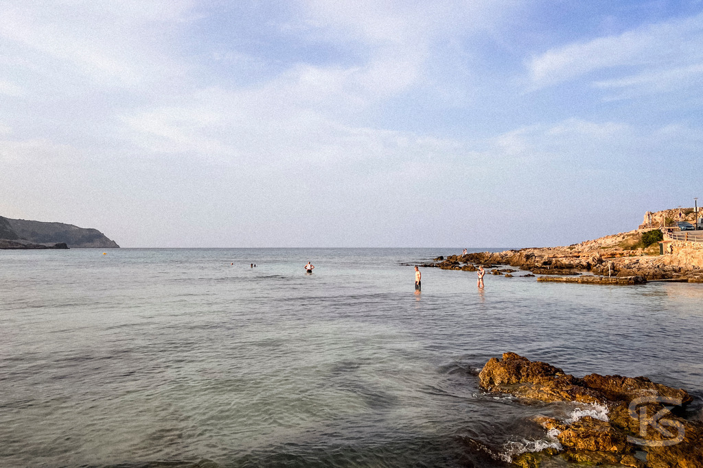 Abendstimmung an der Cala Agulla – Mediterranes Wandbild für Ihr Zuhause | Weiter Blick über das flache Wasser der Bucht Cala Agulla auf Mallorca während des Sonnenuntergangs, mit fernen Personen im Meer und felsiger Küste unter einem hellen Abendhimmel. - Realisiert mit Pictrs.com