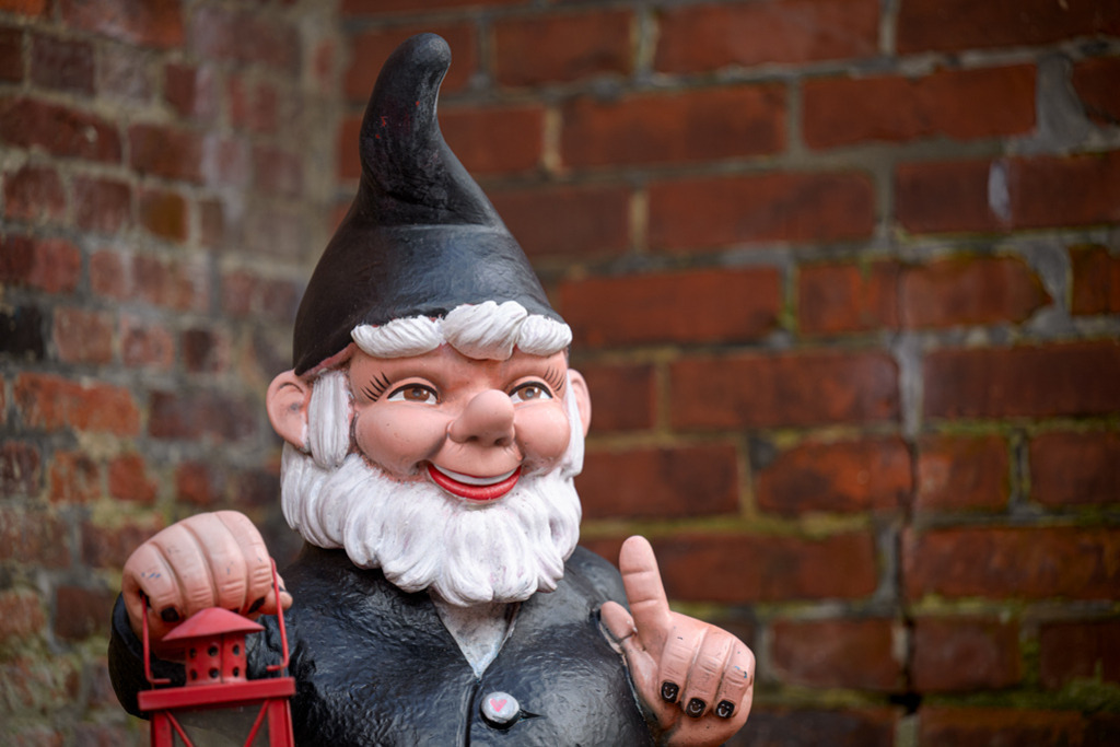 Schwarzer Gartenzwerg | Der freundlich lächelnde Gartenzwerg im schwarzen Gewand wirkt sehr plastisch und nahbar, was durch die unscharfen, tiefroten Ziegel im Hintergrund verstärkt wird. — Auflösung des Originals: 8256 x 5504 px. - Realisiert mit Pictrs.com