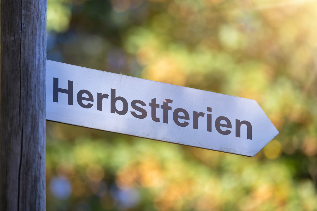 HERBSTFERIEN Schild | (Fotomontage) Richtungspfeil mit der Aufschrift HERBSTFERIEN vor Bäumen mit Herbstfärbung - Realisiert mit Pictrs.com