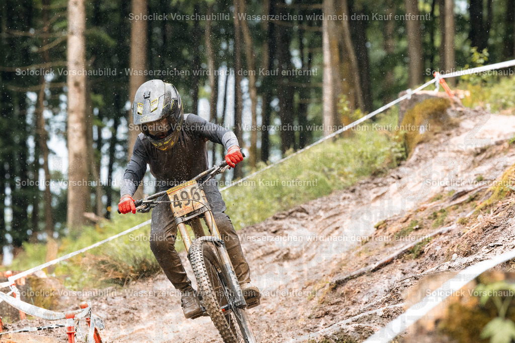 Mountainbike-Action im Wald | Ein Mountainbiker in voller Ausrüstung rast einen staubigen, schmalen Pfad in einem dichten Wald hinunter. Er trägt einen schwarzen Helm mit orangefarbenen Akzenten und eine dunkle Jacke. Er hält ein Mountainbike mit roten Handgriffen fest. Der Pfad ist mit Markierungen versehen, die wahrscheinlich Teil eines Rennens sind. Der Wald ist dicht bewachsen und die Atmosphäre ist dynamisch und actionreich. - Realisiert mit Pictrs.com