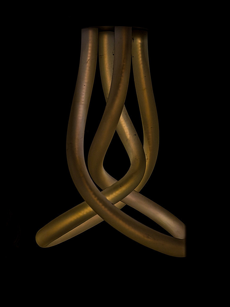 Goldene Symbiose: Abstraktes Licht-Relief vor Tiefschwarz | Ein faszinierendes Meisterwerk minimalistischer Abstraktion. Diese makellose Fine-Art-Fotografie fängt das innere Design einer modernen Leuchtenskulptur ein, deren organisch verschlungene Linien wie flüssiges Gold vor einem abgründigen, tiefschwarzen Hintergrund schweben. Das warme, bronzene Leuchten erzeugt eine beruhigende, fast meditative Tiefe und strahlt puren Luxus aus. Ein exklusives Statement-Piece für modernes High-End-Interieur, Lofts, repräsentative Büros oder das minimalistische Wohnzimmer. Entfaltet seine atemberaubende Eleganz und edle Materialwirkung am besten als glänzender Acrylglas-Print oder als seidenmatter Alu-Dibond-Druck. - Realisiert mit Pictrs.com