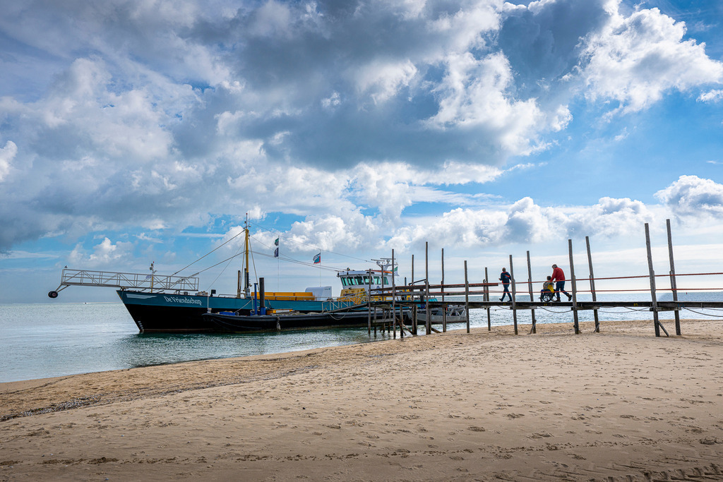 Passagierschiff „De Vriendschap“ am Holzsteg bei De Cocksdorp auf Texel | Das Passagierschiff „De Vriendschap“ liegt am charakteristischen Holzsteg im Norden der Nordseeinsel Texel vor Anker. Personen begehen die Holzbrücke über den Sandstrand am Wattenmeer bei dynamischer Bewölkung. Die Fähre verbindet Texel mit der Nachbarinsel Vlieland und steht symbolisch für maritimen Tourismus, Ausflugsverkehr und barrierefreie Mobilität im Urlaub. - Realisiert mit Pictrs.com