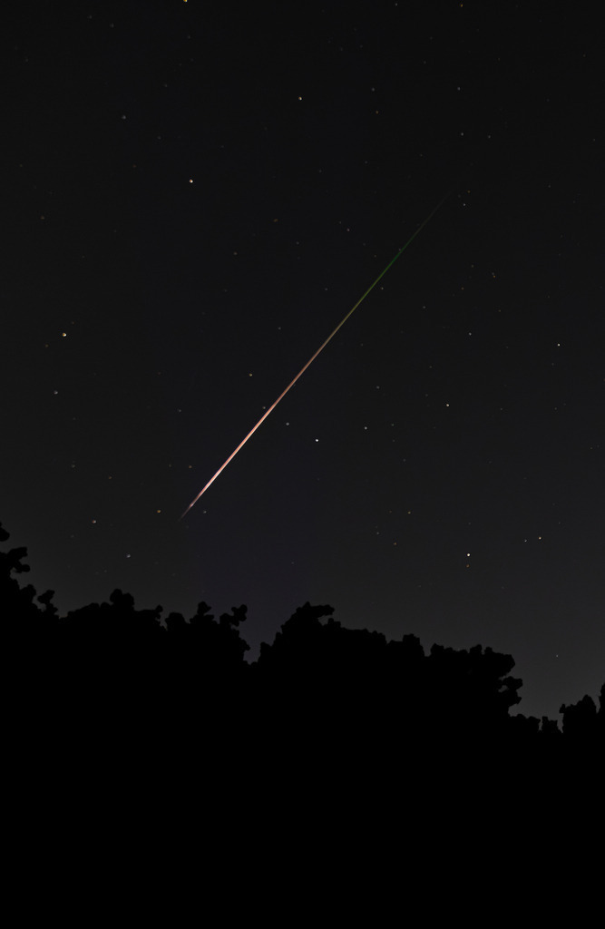 Perseid – A Moment in the Night | Ein Perseiden-Meteor zieht als leuchtende Spur durch den nächtlichen Sternenhimmel. Die Aufnahme entstand am 12. August 2026 während des Perseidenschauers, nahe dem jährlichen Aktivitätsmaximum. Über den dunklen Silhouetten der Bäume zeichnet sich die farbige Leuchtspur deutlich vom tiefen Nachthimmel ab.Ein flüchtiger Augenblick, der nur wenige Sekundenbruchteile sichtbar war – festgehalten als atmosphärische Aufnahme zwischen Astrofotografie, Natur und Fine Art. - Realisiert mit Pictrs.com