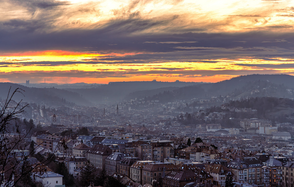 Sonnenuntergang über der Stadt Stuttgart-Talkessel-Häuser | Das Bild zeigt einen Panoramablick auf die Stadt Stuttgart in Deutschland bei Sonnenuntergang oder Sonnenaufgang. Die Aufnahme wurde wahrscheinlich von einem Hügel aus gemacht, möglicherweise der Karlshöhe, um die weite Sicht über das Tal zu ermöglichen.Die Atmosphäre ist durch eine Dunstschicht über der Stadt und dramatische, goldgelbe und dunkle Wolken am Himmel geprägt.Das Bild trägt ein Wasserzeichen mit dem Text "tom-pic-art.de".Die Beleuchtung lässt auf die "Blaue Stunde" oder "Goldene Stunde" schließen, was für stimmungsvolle Lichtverhältnisse sorgt. - Realisiert mit Pictrs.com
