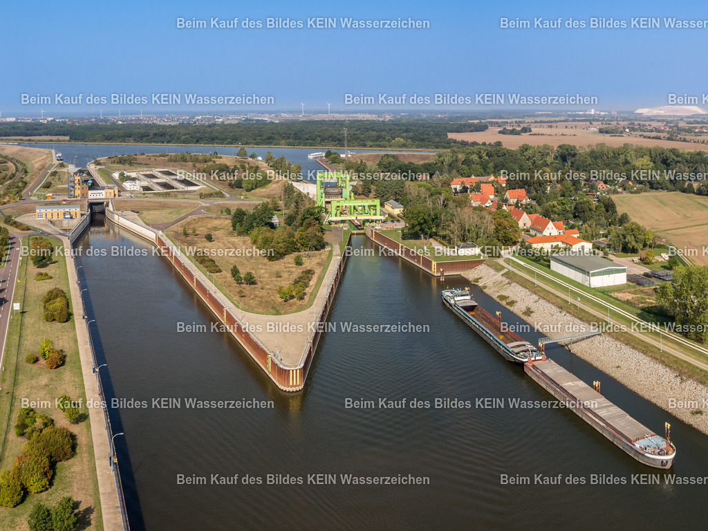 Schiffshebewerk-2 | Das Luftbild zeigt das Schiffshebewerk Rothensee und die benachbarte Doppelsparschleuse Hohenwarthe an einem sonnigen Sommertag aus einer Drohnenperspektive. Die beiden Wasserbauwerke bilden gemeinsam mit der Trogbrücke das Wasserstraßenkreuz Magdeburg – einen der bedeutendsten Verkehrsknotenpunkte der europäischen Binnenschifffahrt. Aus der Vogelperspektive werden die Dimensionen und die unterschiedlichen technischen Konzepte beider Anlagen eindrucksvoll sichtbar.Das Schiffshebewerk Rothensee wurde 1938 in Betrieb genommen und überwindet einen Höhenunterschied von 16 Metern mithilfe eines 5.400 Tonnen schweren, wassergefüllten Troges. Die Doppelsparschleuse Hohenwarthe ergänzt die Anlage seit 2003. Ihre beiden Schleusenkammern sind jeweils 190 Meter lang und 12,50 Meter breit. Mit einer Fallhöhe von bis zu 19 Metern und acht Sparbecken werden bei jedem Schleusenvorgang bis zu 60 Prozent des benötigten Wassers eingespart – ein wesentlicher Vorteil gegenüber herkömmlichen Schleusen.Die Drohnenaufnahme dokumentiert eindrucksvoll das Zusammenspiel historischer und moderner Wasserbautechnik. Schiffshebewerk und Doppelsparschleuse stehen beispielhaft für mehr als 80 Jahre Ingenieurentwicklung und machen das Wasserstraßenkreuz Magdeburg zu einem der leistungsfähigsten Binnenverkehrsknoten Europas. - Realisiert mit Pictrs.com