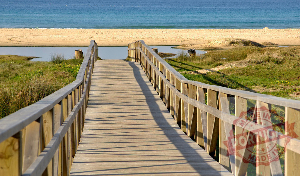 Wooden boardwalk leading to a sandy beach and blue ocean. Espana - Cadiz | Wooden boardwalk crossing wetlands to a sunny sandy beach and blue ocean - Realisiert mit Pictrs.com