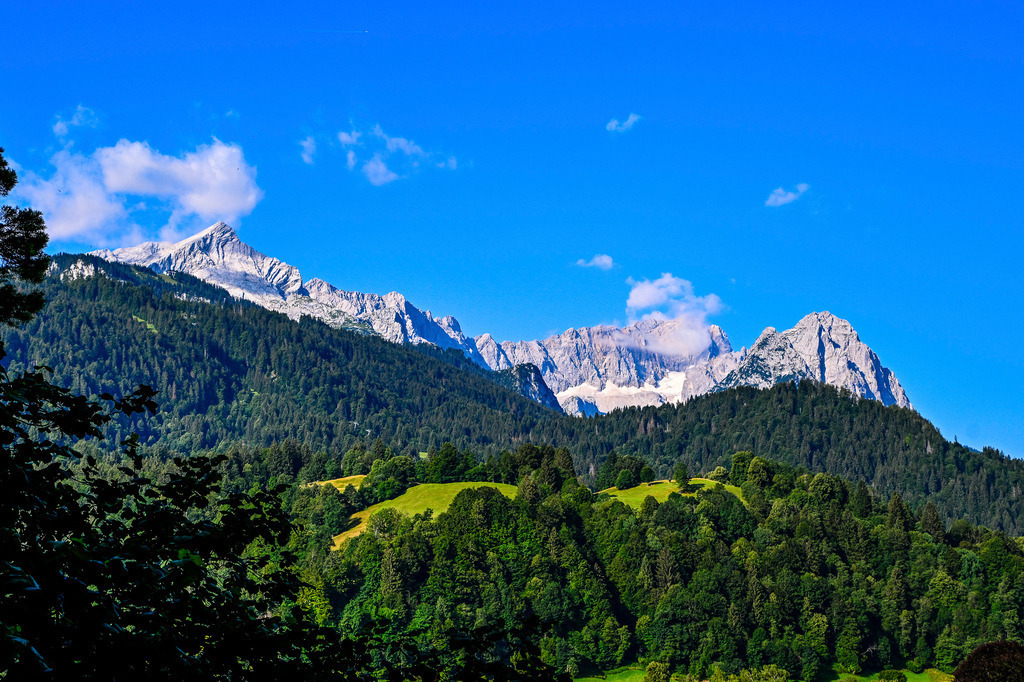 Sommerliche Alpen: Von der Alpspitze zur Zugspitze | Ein Sommertraum in sattem Grün und strahlendem Blau: Das majestätische Wettersteingebirge zeigt sich in seiner vollen, erhabenen Pracht. Links grüßt die markante Pyramide der Alpspitze, während der Blick über die schroffe Bergkette bis hin zur mächtigen Zugspitze schweift, deren Flanken von sanften Wolkenschleiern umspielt werden. Im Vordergrund leuchten die saftigen, sonnenbeschienenen Almwiesen inmitten dichter Bergwälder. Diese Aufnahme, festgehalten im Juli im Umland von Garmisch-Partenkirchen, versprüht pure Lebensfreude, die Sehnsucht nach alpiner Freiheit und die tiefe Ruhe eines perfekten Sommertages in den Bergen. - Realisiert mit Pictrs.com
