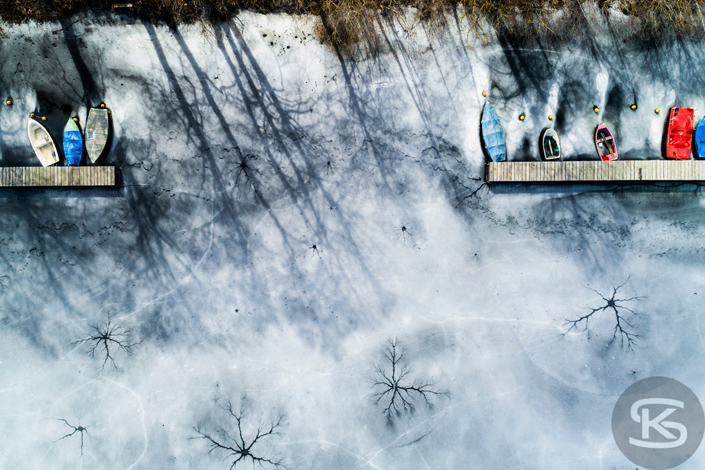 Eingefrorene Boote am Wintersteg – Drohnenaufnahme eines zugefrorenen Sees | Beeindruckende Luftaufnahme von eingefrorenen Booten am Steg eines zugefrorenen Sees. Die Drohnenfotografie zeigt winterliche Stille mit Baumschatten auf der Eisfläche aus der Vogelperspektive – atmosphärische Winterimpression. - Realisiert mit Pictrs.com