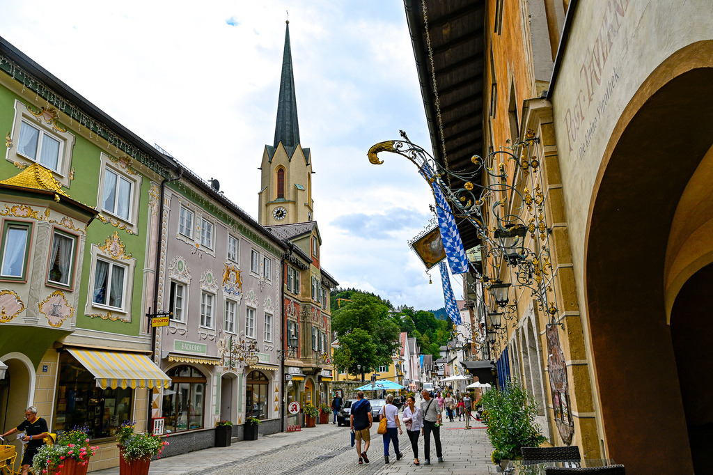 Geschichte und Charme der Partenkirchener Ludwigstraße | Bunte, kunstvoll verzierte Fassaden mit traditioneller Lüftlmalerei, schmiedeeiserne Nasenschilder und der spitz aufragende Kirchturm von Maria Himmelfahrt: Die historische Ludwigstraße in Partenkirchen offenbart ihr charmantestes Gesicht. Beim gemütlichen Bummeln über das Pflaster spürt man den lebendigen Atem bayerischer Geschichte, gepaart mit moderner Gastfreundschaft und alpenländischer Gemütlichkeit – ein zeitloser Ort, der zum Verweilen, Staunen und Genießen einlädt. - Realisiert mit Pictrs.com