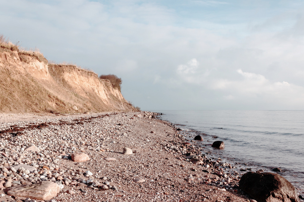 Wandbild: Steilküste am Meer in dezenten Farben | Dieses Wandbild zeigt die Steilküste am Meer in dezenten Farben. Die Abbruchkante der Steilküste ist beige und braun. Zwischen der Steilküste und dem Meer befindet sich ein Naturstrand an dem viele Steine liegen. Schaffen Sie sich ein maritimes Ambiente in Ihrem Wohnzimmer und holen Sie sich dieses stilvolle Wandbild. Es ist auf Leinwand, Aluminium-Platte, Acrylglas oder als Holzdruck erhältlich. Die Wandbilder werden individuell für Sie in vielen Abmessungen produziert. Daher passen die Ostseekult Wandbilder immer perfekt an Ihre Wände. - Realisiert mit Pictrs.com