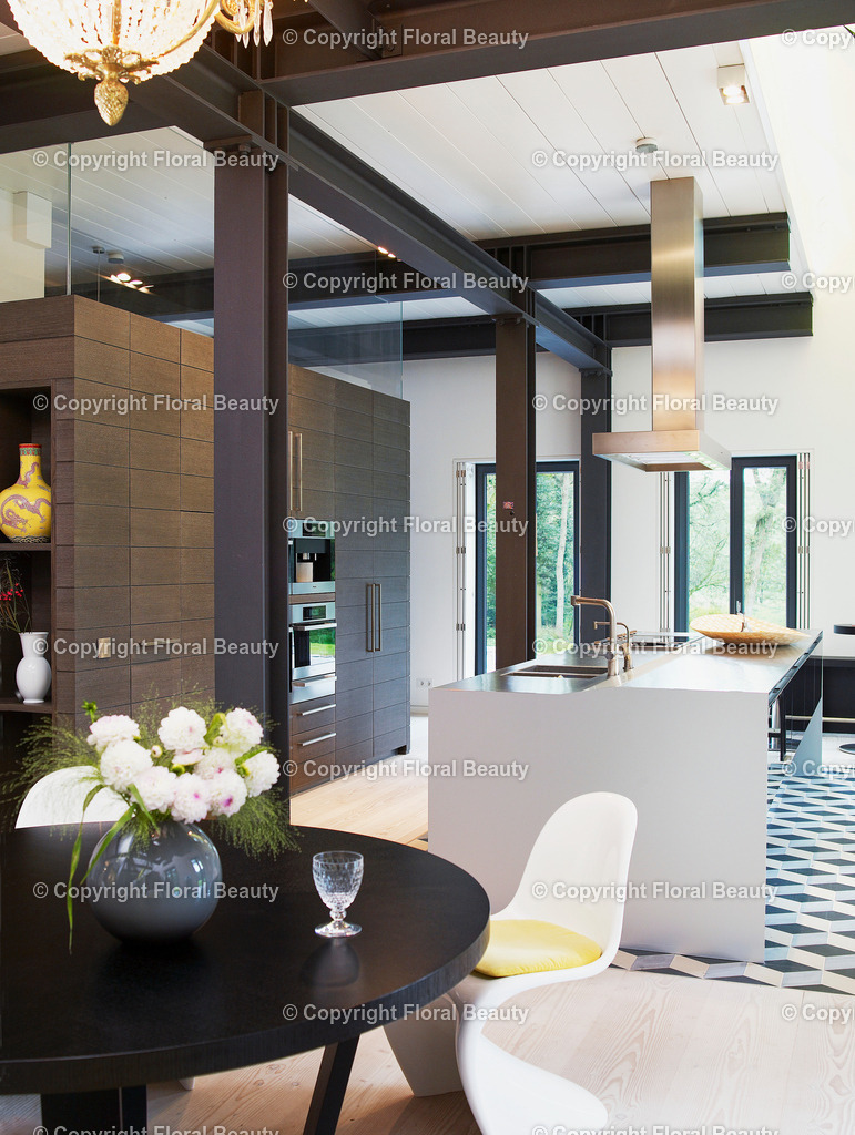Moderne Küche in schwarz-weiß-grau | Offene Küche mit  Edelstahl Kochinsel. Küchenschränke aus dunkler Holzvertäfelung. Waschbetonfliesen in grau-schwarz-beige, Rautenform und weißer Dielenboden. Im Vordergrund schwarzer Esstisch mit weißen Panton Stühlen. - Realisiert mit Pictrs.com