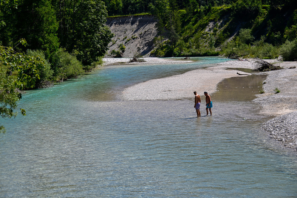 Oase der Stille: Sommerglück an der wilden Isar | Ein Anblick voller Leichtigkeit, Entschleunigung und sommerlicher Sehnsucht: Kristallklar und in leuchtenden Türkistönen schlängelt sich die ungezähmte Isar durch ihr naturbelassenes Tal. Auf einer der weiten, hellen Kiesbänke stehen zwei Menschen knöcheltief im erfrischenden Wasser und genießen den Moment der Ruhe. Eingerahmt von saftig grünen Ufern und einer markanten Steilwand im Hintergrund verströmt diese Szenerie die pure Harmonie eines Sommertages. Diese Aufnahme fängt die magische Verbindung zwischen Mensch und unberührter Natur perfekt ein – ein Bild, das zum Verweilen einlädt und die Seele atmen lässt. - Realisiert mit Pictrs.com