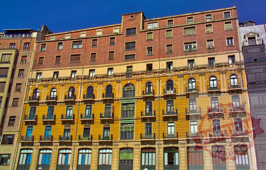 City building facade with ornate balconies and colorful windows. Spain - Bilbao | Building facade featuring architectural details, arched windows, and small balconies under a blue sky - Realisiert mit Pictrs.com