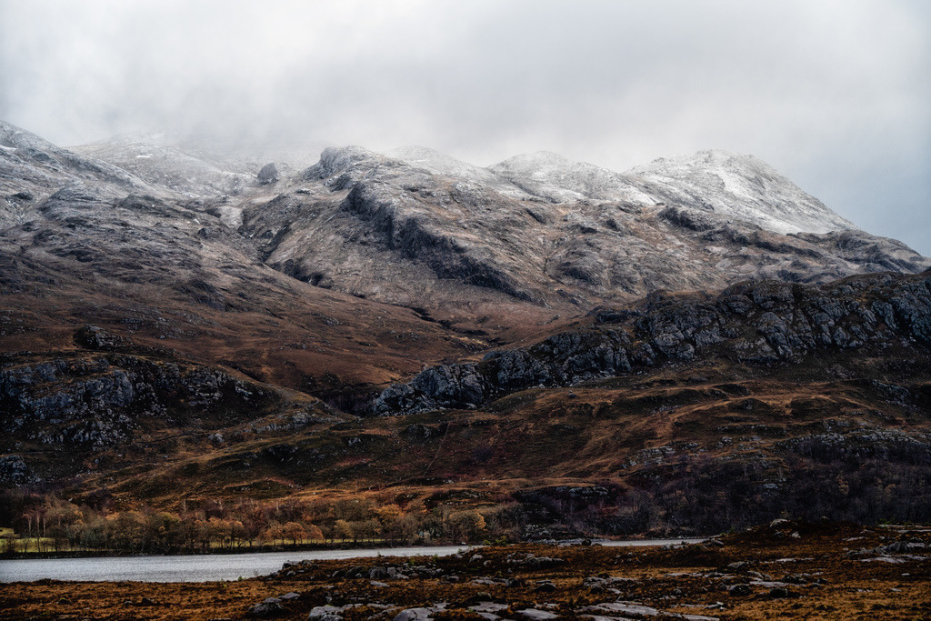 Winterlandschaft am Loch Maree | Eine weite Berglandschaft mit schneebedeckten Gipfeln und einem See erstreckt sich unter einem bewölkten Himmel. Die unteren Hänge zeigen eine Mischung aus felsigen Strukturen und herbstlicher Vegetation in Brauntönen. Der ruhige Loch Maree bildet einen Kontrast zu den rauen, nebelverhangenen Bergen. - Realisiert mit Pictrs.com