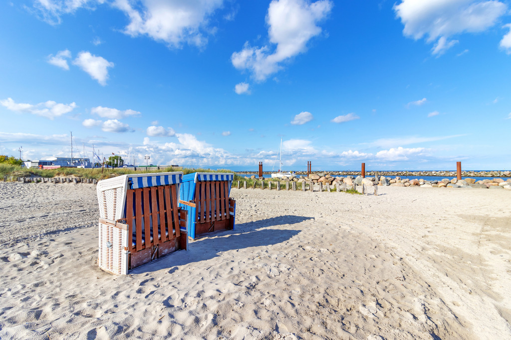 XXL Wandbild: Zwei Strandkörbe am Meer | Dieses Wandbild im Querformat zeigt zwei Standkörbe am Sandstrand. Im Hintergrund ist eine Steinmole zu sehen. Am schönen blauen Himmel befinden sich einige Schäfchenwolken. - Realisiert mit Pictrs.com