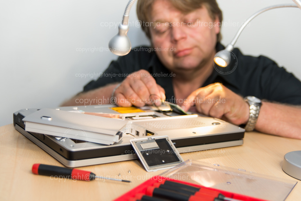 Mann repariert Laptop  Notebook D | Ein Gehäuse eines Notebooks/Laptops ist geöffnet. Zwei Männerhände stecken einen neuen Speicher in den Schlitz. 2 Lampen und feine Werkzeuge.
