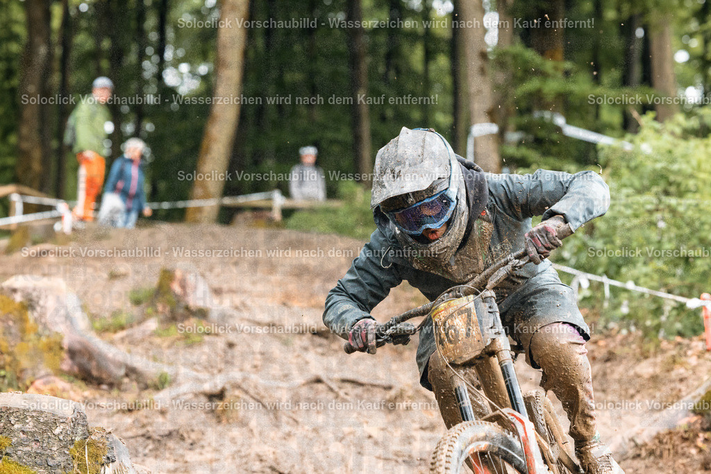 Mountainbike-Action im Wald | Ein Mountainbiker, bedeckt mit Schlamm, fährt mit hoher Geschwindigkeit einen steilen, schlammigen Trail in einem dichten Wald hinab. Er trägt einen Helm, eine Schutzbrille und spezielle Mountainbike-Bekleidung. Der Trail ist mit Wurzeln und Steinen übersät. Im Hintergrund sind Zuschauer zu sehen, die den Wettkampf beobachten. Die Szene vermittelt ein Gefühl von Geschwindigkeit, Abenteuer und Herausforderung. - Realisiert mit Pictrs.com