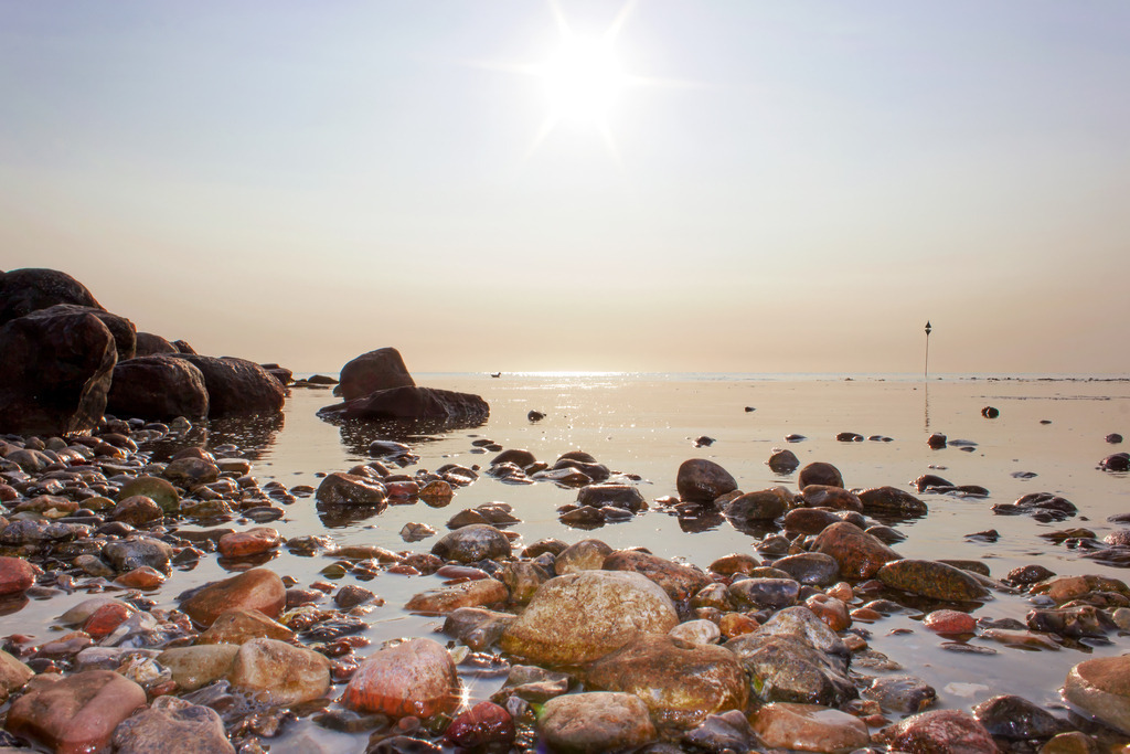 Wandbild: Sanftes Erwachen am Naturstrand | Ein Erwachen voller Glanz und Stille. Dieses Wandbild fängt den Moment ein, in dem die aufgehende Sonne einen Steinstrand an der Ostsee in ein Meer aus Licht taucht. Während die massive Steinmole im Hintergrund für Ruhe und Beständigkeit sorgt, funkeln die nassen Kiesel im Vordergrund wie kleine, geschliffene Edelsteine. Eine haptische und zugleich meditative Komposition, die die reine, klare Morgenluft des Nordens direkt in Ihre Räume bringt. - Realized with Pictrs.com