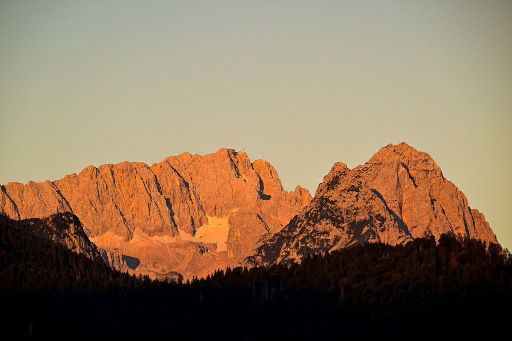 Glühende Giganten: Das erste Licht über der Zugspitze | Ein Anblick von erhabener Stille und magischer Herzenswärme: Im sanften Schein der Morgensonne erstrahlt das mächtige Massiv der Zugspitze und der angrenzenden Gipfel in einem warmen, intensiven Alpenglühen. Die schroffen, zerfurchten Felswände leuchten in sanften Gold- und Orangetönen, während tief in den Karen letzte Schneefelder hell hervorstechen. Am höchsten Punkt zeichnet sich fein die Silhouette der Bergstation ab. Im krassen Kontrast dazu liegt der dichte Nadelwald im Vordergrund noch in tiefem, mystischem Schatten. Diese Aufnahme fängt den flüchtigen, friedlichen Moment des erwachenden Tages beim Sonnenaufgang ein – ein Bild voller Harmonie und zeitloser Geborgenheit im Hochgebirge. - Realisiert mit Pictrs.com