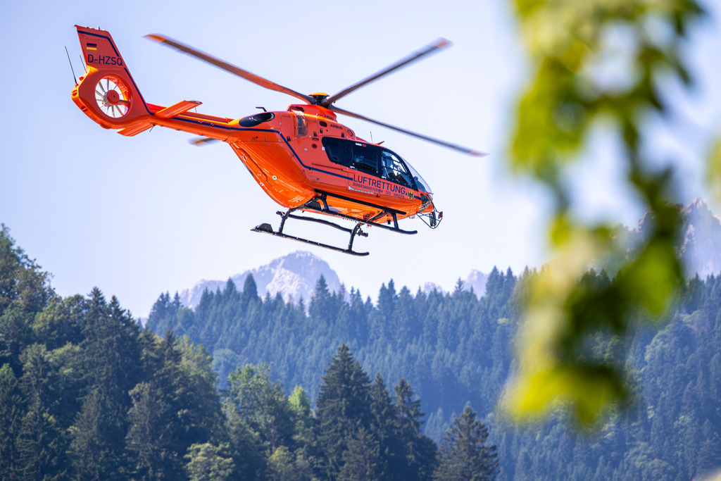 Rettungshubschrauber | Der orangefarbene Hubschrauber (D-HZSQ) fliegt in einer dynamischen Kurve vor der imposanten Kulisse bewaldeter Berghänge und schroffer Felsgipfel. Ein unscharfer grüner Zweig im Vordergrund verleiht der Szene Tiefe und rahmt den Flugmoment natürlich ein. - Realisiert mit Pictrs.com