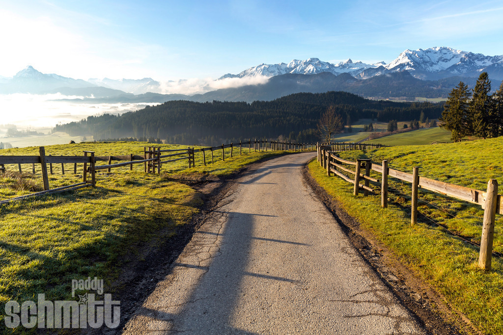 Der Weg ins Allgäu | Ein e Landstraße führt durch die idyllische Landschaft des Allgäus. Umgeben von grünen Wiesen und sanften Hügeln und fantastischer Bergsicht, lädt die Natur zum Entdecken und Genießen ein. - Realisiert mit Pictrs.com
