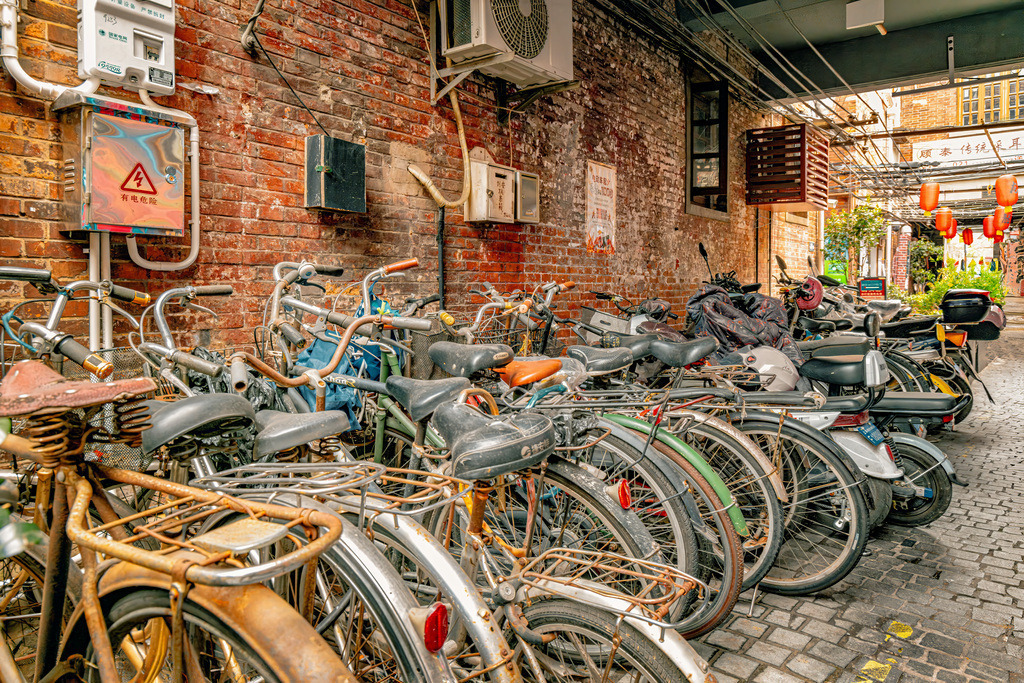 0Y9A8894-Bearbeitet Kopie | Das ausgewählte Bild zeigt eine enge, traditionelle Gasse in China, die dicht mit Fahrrädern und Motorrollern vollgestellt ist. Die Backsteinwände, die freiliegenden Kabelstränge, Klimaanlagen an den Wänden sowie die roten Schilder im Hintergrund sind charakteristisch für traditionelle Wohnviertel in chinesischen Großstädten (oft als Hutongs in Peking oder als Gassen in Tianzifang, Shanghai, zu finden).Es zeigt dichte Reihen älterer, klassischer Fahrräder neben moderneren E-Bikes - Realisiert mit Pictrs.com