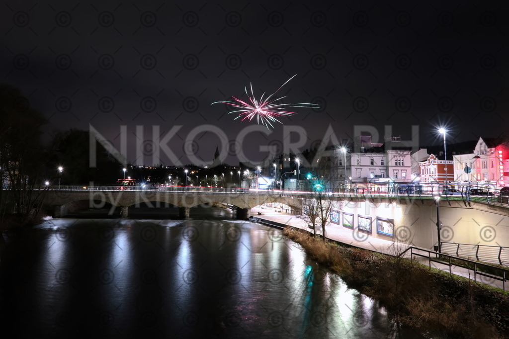 Nachtaufnahme der Lennepromenade in Letmathe | Die Lennepromenade in Letmathe bei Nacht. Die Laternenlichter der Lennebrücke spiegeln sich in der Lenne. Über der Brücke erleuchtet eine Feuerwerksrakete den Nachthimmel.