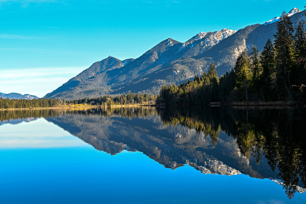 Spiegel der Einsamkeit: Die vollkommene Stille am Barmsee | Ein Anblick von magischer Ruhe und tiefer Klarheit, der die Seele sanft umarmt: Wie ein makelloser, tiefblauer Spiegel liegt der Barmsee in der winterlichen Bergwelt. Die majestätischen, teils schneebedeckten Hänge des Karwendelgebirges und die dichten, dunkelgrünen Nadelwälder spiegeln sich in perfekter Symmetrie auf der kristallklaren Wasseroberfläche. Keine Welle stört dieses friedliche Kunstwerk der Natur unter dem strahlend blauen Himmel. Diese Aufnahme fängt einen kostbaren Moment der absoluten Windstille und Einsamkeit ein – ein Ort des Rückzugs, der zum Träumen und tiefen Durchatmen einlädt. - Realisiert mit Pictrs.com