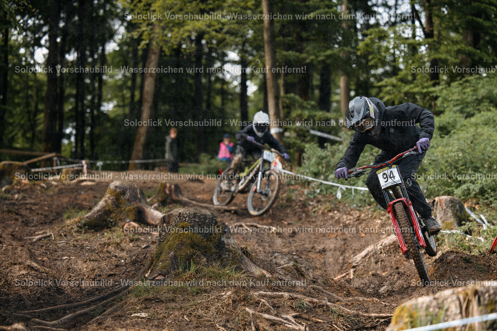 Mountainbike-Action im Wald | Das Foto zeigt einen Mountainbiker in voller Ausrüstung, der mit hoher Geschwindigkeit einen staubigen, kurvigen Kurs durch ein dichtes Waldgebiet durchquert. Er trägt einen schwarzen Helm mit einer Visier und eine dunkle Jacke mit dem Nummernschild '94'. Im Hintergrund ist ein weiterer Mountainbiker zu sehen, der ebenfalls den Kurs befährt. Der Kurs ist mit Baumstümpfen und Wurzeln übersät, was die Herausforderung für die Fahrer unterstreicht. - Realisiert mit Pictrs.com