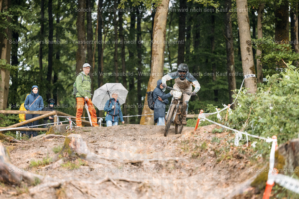 Mountainbike-Action im Wald | Ein Mountainbiker in voller Montur, inklusive Helm und Schutzbrille, rast einen staubigen, kurvigen Trail in einem dichten Wald hinunter. Der Trail ist mit Baumstümpfen und Wurzeln übersät. Mehrere Zuschauer stehen am Rand des Trails und beobachten das Rennen. Einige halten Regenschirme, was auf feuchtes Wetter hindeutet. Die Szene vermittelt ein Gefühl von Action und Abenteuer. - Realisiert mit Pictrs.com