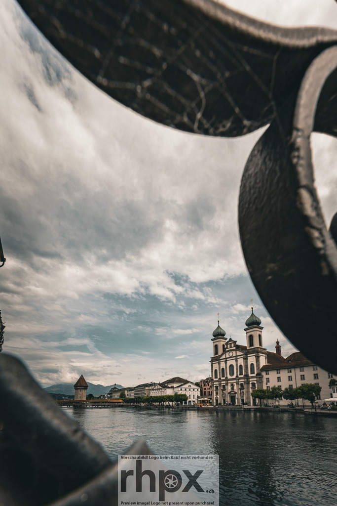 Jesuitenkirche und Kapellbrücke hinter Ornamenten | Rahmen. Barock. Flusslauf.Ein kunstvoller Blick durch ein verschnörkeltes Detail auf die markante Silhouette der Jesuitenkirche am Reussufer. Die architektonische Pracht wird durch die spielerische Rahmung im Vordergrund in eine ganz neue, faszinierende Perspektive gerückt.Wähle unter "Produktauswahl" dein Wunschformat: Vom klassischen Wandbild über Puzzle &amp; Tassen bis zum digitalen Download (z.B. als Handy-Hintergrund). - Realisiert mit Pictrs.com