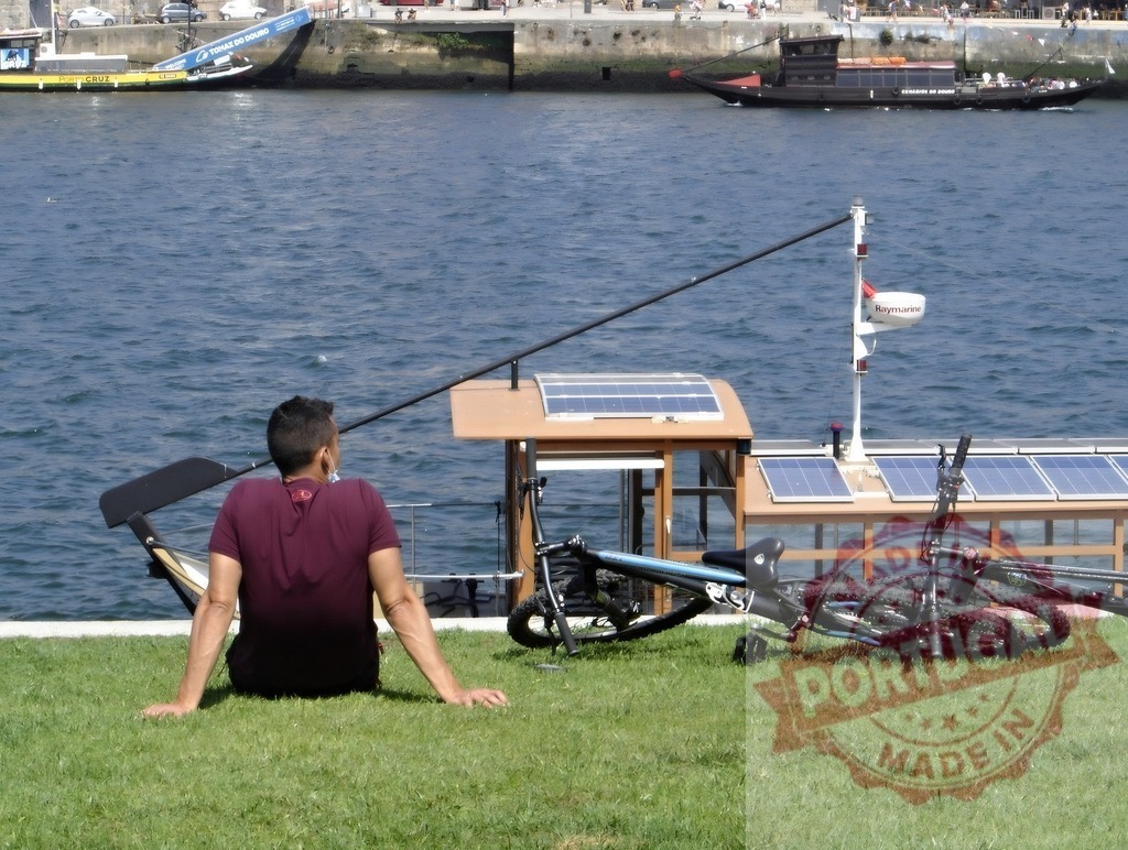 Man relaxing by Douro river in Porto with bike and solar panels. Porto - Portugal | Man relaxing on grass near river, a bicycle and solar powered boat at a dock - Realized with Pictrs.com