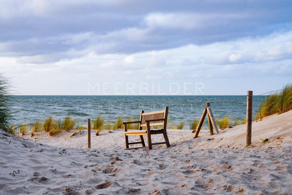Ahrenshoop Ostsee Bilder II | Eine Bank am Dünenweg in Ahrenshoop wurde hier aus dem Schatten heraus fotografiert. Dadurch erstrahlt die Bank sanft im letzten Sonnenlicht des Tages. Im Hintergrund erstrecken sich leichte Dünen, die scheinbar nahtlos in die Ostsee übergehen. Doch zwischen den Dünen und der Ostsee liegt der Strand. Der vordere Teil des Bildes ist absichtlich leicht unscharf gehalten. Dieses wunderschöne Motiv an der Ostsee entfaltet seine volle Wirkung besonders auf einer klassischen Leinwand oder einem Alu-Dibond-Druck.