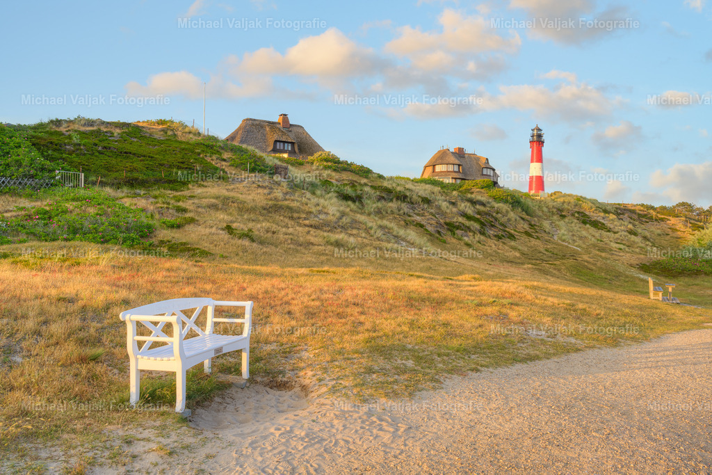 Weiße Bank in Hörnum | Eine weiße Bank in Hörnum im Süden von Sylt lädt zu einer Rast ein. Im Hintergrund sind Reetdachhäuser und der Hörnumer Leuchtturm zu sehen. - Realisiert mit Pictrs.com