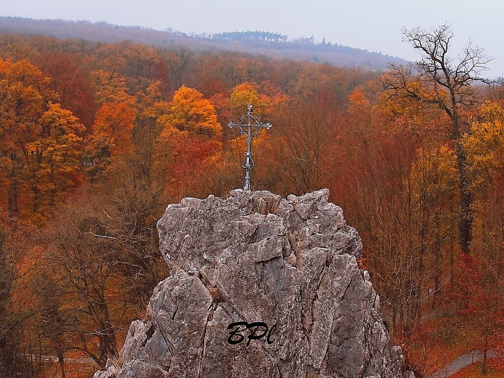 Bergkreuz im Herbst | Auf dem Bild ist im Vordergrund ein Bergkreuz zu sehen. Der Hintergrund erschließt sich aus der roten Farbenpracht der Herbstbäume. durch den Farbunterschied zwischen dem Berg und den Blättern entsteht ein starker Kontrast. - Realisiert mit Pictrs.com