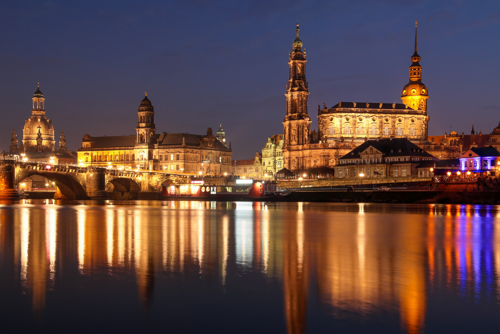 Abend an der Elbe - Dresdens historische Silhouette | Dieses Panorama-Wandbild zeigt die Dresdner Altstadt in ihrer ganzen Pracht. Im warmen Licht der Nacht erstrahlen die Frauenkirche, die Hofkirche und das Residenzschloss entlang der Elbe. Die beleuchtete Augustusbrücke spannt sich elegant über das ruhige Wasser, das die nächtliche Szene eindrucksvoll spiegelt. Ein harmonisches Zusammenspiel aus Geschichte, Architektur und Licht. Ideal für alle, die stilvolle Stadtansichten und eine ruhige, elegante Bildsprache schätzen.