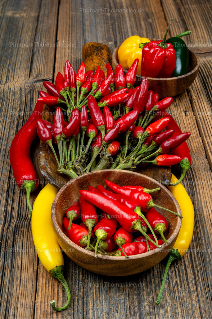 Bunte scharfe Chilis in Holzschalen auf altem Holztisch | Auf einem rustikalen Holztisch präsentieren sich verschiedene Chilisorten und Paprika in natürlichen Holzschalen. Leuchtend rote Chilischoten dominieren eine große Schale, während bunte Paprika in Rot, Gelb und Grün eine zweite Schale füllen. Die kleinen scharfen Chilis mit ihren grünen Stielen bilden ein lebendiges Arrangement auf der warmen Holzoberfläche. Diese harmonische Komposition zeigt die Vielfalt scharfer Küchengewürze in ihrer natürlichen Schönheit. Die verschiedenen Formen und Farben schaffen eine appetitliche Darstellung hochwertiger Zutaten. - Realisiert mit Pictrs.com