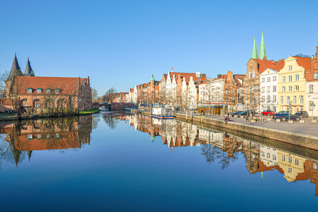 Lübeck – An der Obertrave | Die Trave in Lübeck grenzt die historische Altstadt nach Westen ab. In der Fotografie befinden sich auf der linken Seite das Holstentor und die Salzspeicher, rechts erhebt sich die Häuserzeile der Straße: An der Obertrave. — Auflösung des Originals: 5982 x 3988 px. - Realisiert mit Pictrs.com