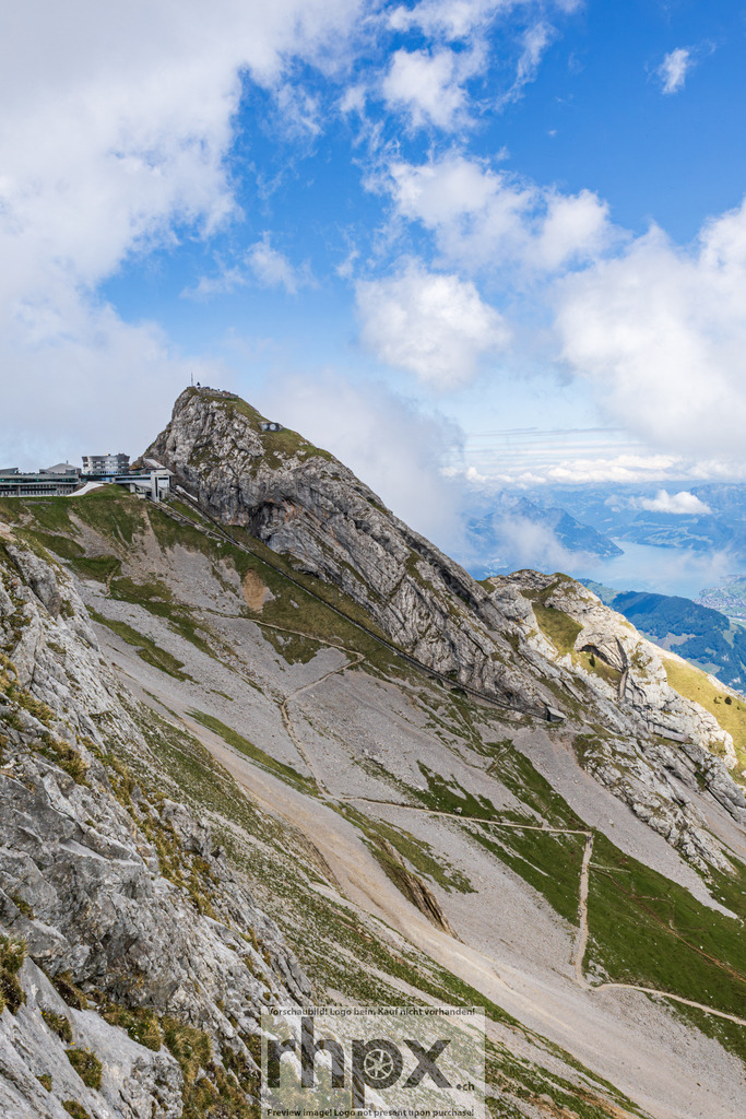 Pilatus Gipfelgrat mit Wanderweg, Vierwaldstättersee und Tal | Blick vom Grat des Pilatus auf den Hotelkomplex Pilatus-Kulm (Eselsgipfel, 2132 m ü. M.) links oben. Im Mittelgrund sichtbarer Wanderweg auf dem Grashang. Im Hintergrund Vierwaldstättersee mit Ortschaft und grünem Alpental. - Realisiert mit Pictrs.com