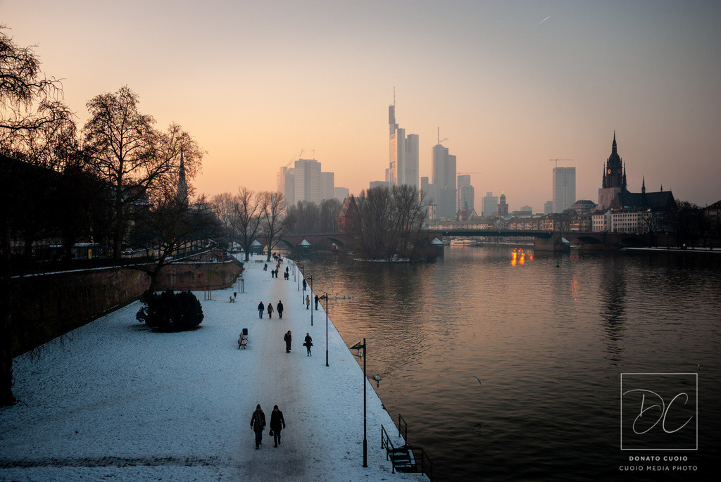 Frankfurt – Winter am Main | Ein seltenes Winterbild von Frankfurt: klare Kälte, warme Abendfarben, ruhige Spaziergänger am verschneiten Mainufer. Dieser Fine-Art-Print verbindet urbanes Leben mit poetischer Stille und einer leichten Melancholie, die besonders im Winter in der Stadt liegt.Frankfurt Skyline im Winter bei Sonnenuntergang mit Schnee und Spaziergängern am Main - Realisiert mit Pictrs.com