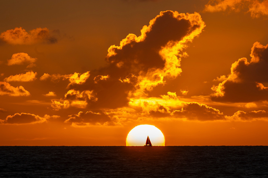 Dramatische Abendstimmung über der Nordsee | Ein Segelboot zeichnet sich als feine Silhouette direkt vor der leuchtend gelben Scheibe der untergehenden Sonne ab, die am flachen Horizont der Nordsee versinkt. Über der Küste der niederländischen Insel Texel erstreckt sich ein von warmen Orange- und Goldtönen durchzogener Wolkenhimmel. Eine stimmungsvolle Aufnahme für redaktionelle Beiträge zu den Themen Segelsport, Seefahrt, Nordseeurlaub, Naturromantik und sommerliche Abendstimmungen. - Realisiert mit Pictrs.com
