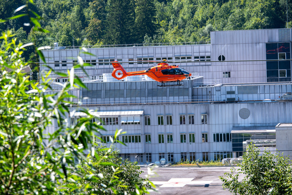 Rettungshubschrauber | Ein leuchtend oranger Rettungshubschrauber der Zivilschutz-Luftrettung schwebt im Anflug über dem modernen, weitläufigen Klinikum Garmisch-Partenkirchen. Während die technische Kühle der silbernen Fassade den Hintergrund dominiert, setzen grüne Blätter im Vordergrund einen lebendigen, natürlichen Akzent und rahmen die Szene ein. - Realisiert mit Pictrs.com