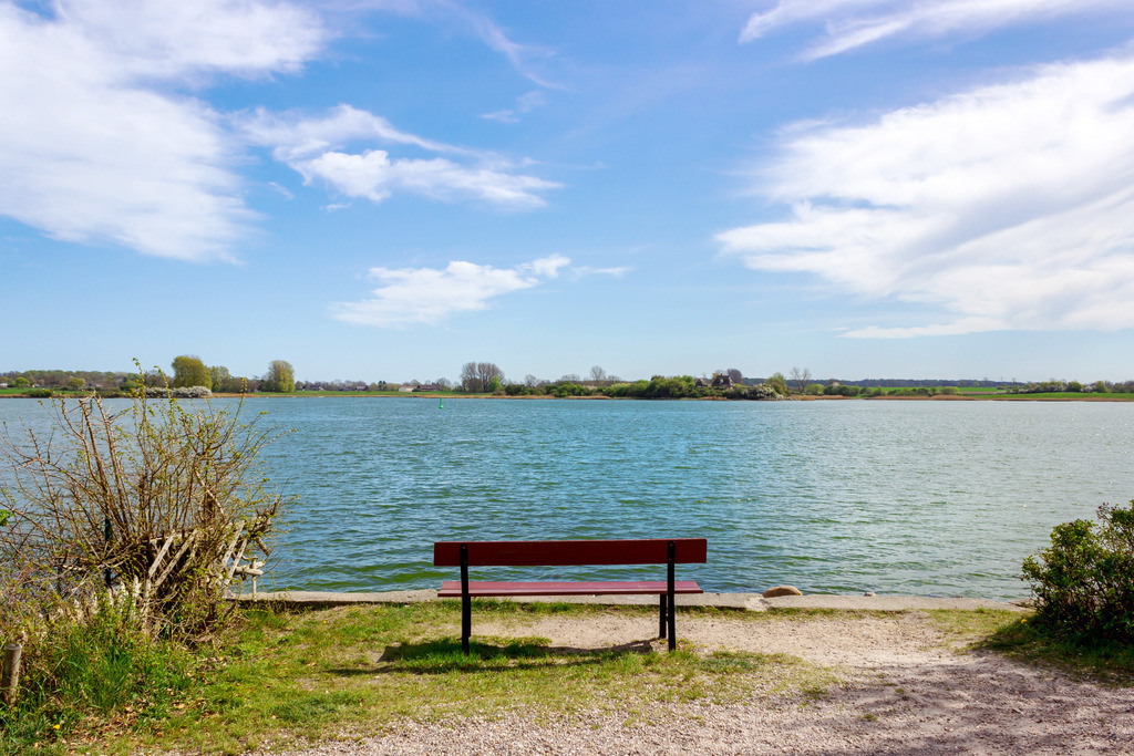 Wandbild: Bank in Arnis an der Schlei  | Dieses Wandbild im Querformat zeigt eine Bank direkt am Schleiufer. Am blauen Himmel sind schöne helle Wolken zu sehen. - Realisiert mit Pictrs.com