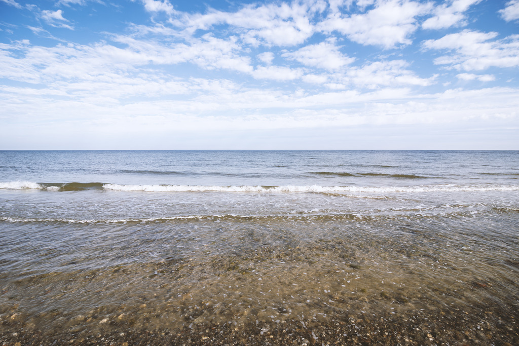 Wandbild: Horizont der Ruhe - Weitblick über Falshöft | Dieses lichtdurchflutete Wandbild bringt die belebende Frische der Ostsee direkt in den Raum. Im Vordergrund gibt das transparente, sanft kräuselnde Wasser den Blick frei auf den feinen Kieselgrund, während sich darüber der weite Horizont unter einem malerischen Wolkenhimmel erstreckt. Eine zeitlos elegante Fotokunst, die Räumen eine natürliche Leichtigkeit, Tiefe und ein Gefühl von unendlicher Freiheit verleiht – perfekt für stilvolle, moderne Interieurs.