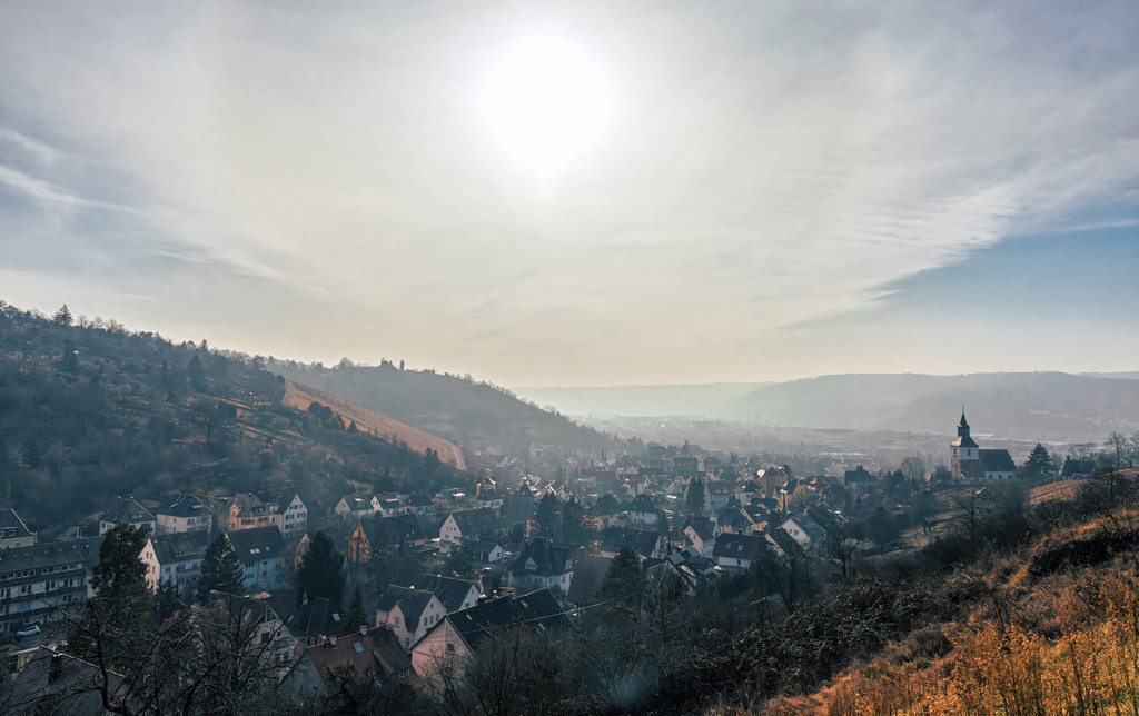 Romantischer Blick über Stuttgart Obertürkheim und seine Weinberge | Das Bild zeigt eine Panoramaaufnahme von Stuttgart Obertürkheim. Die Aufnahme wurde an einem sonnigen Tag von einem erhöhten Aussichtspunkt aus gemacht und zeigt den Ort eingebettet in eine hügelige Landschaft. Die Szene wird durch einen hellen Lichtschein im oberen Zentrum des Bildes geprägt und zeigt eine Mischung aus Wohngebäuden und der charakteristischen Landschaft der Region.  - Realisiert mit Pictrs.com