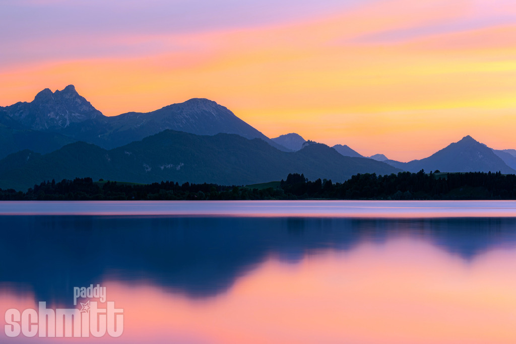 Abendstimmung am Forggensee bei Füssen | Stimmungsvolle Abendstimmung am Forggensee bei Füssen, während das letzte Licht des Tages die Landschaft in warme Farben taucht. Die Berge spiegeln sich sanft im ruhigen Wasser. - Realisiert mit Pictrs.com