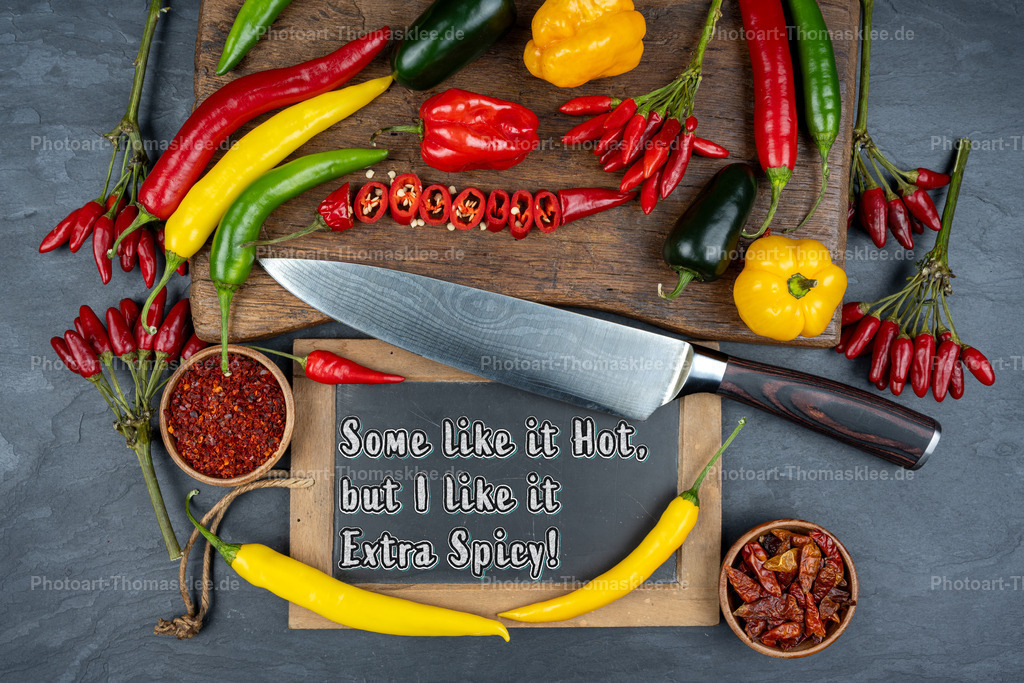 Scharfe Chilis und Gewürze - Extra Spicy Küchenkunst | Auf einem rustikalen Holzbrett liegen verschiedene Chilisorten in leuchtenden Farben. Ein scharfes Küchenmesser ergänzt die Komposition perfekt. Rote, gelbe und grüne Paprika bilden einen farbenfrohen Kranz um eine schwarze Schiefertafel mit dem Spruch "Some like it Hot, but I like it Extra Spicy!". Kleine Schüsseln mit Chilipulver und getrockneten Chilis runden das Arrangement ab. Das Bild strahlt pure Leidenschaft für scharfe Küche aus. - Realisiert mit Pictrs.com