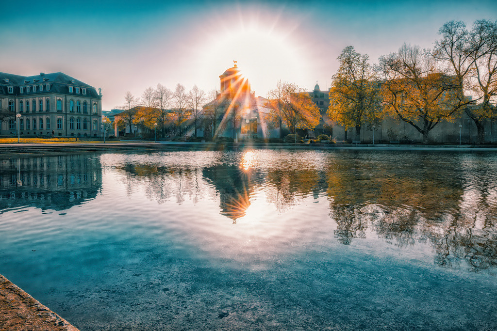 Der strahlende Eckensee in Stuttgart,Landesbibliothek | Das Bild zeigt den Eckensee in Stuttgart, mit dem Gebäude der Württembergischen Landesbibliothek im Hintergrund, aufgenommen bei Sonnenaufgang oder Sonnenuntergang. Der Eckensee ist ein künstlicher See im Herzen Stuttgarts, gelegen zwischen dem Landtag von Baden-Württemberg und der Württembergischen Landesbibliothek.Das im Hintergrund sichtbare Gebäude beherbergt die Württembergische Landesbibliothek, eine der größten wissenschaftlichen Universalbibliotheken in Baden-Württemberg.Die Aufnahme fängt die malerische Reflexion des Gebäudes und der Bäume im ruhigen Wasser des Sees ein.Das intensive Sonnenlicht erzeugt einen strahlenförmigen Lichteffekt, der die Szene dramatisch beleuchtet. - Realisiert mit Pictrs.com