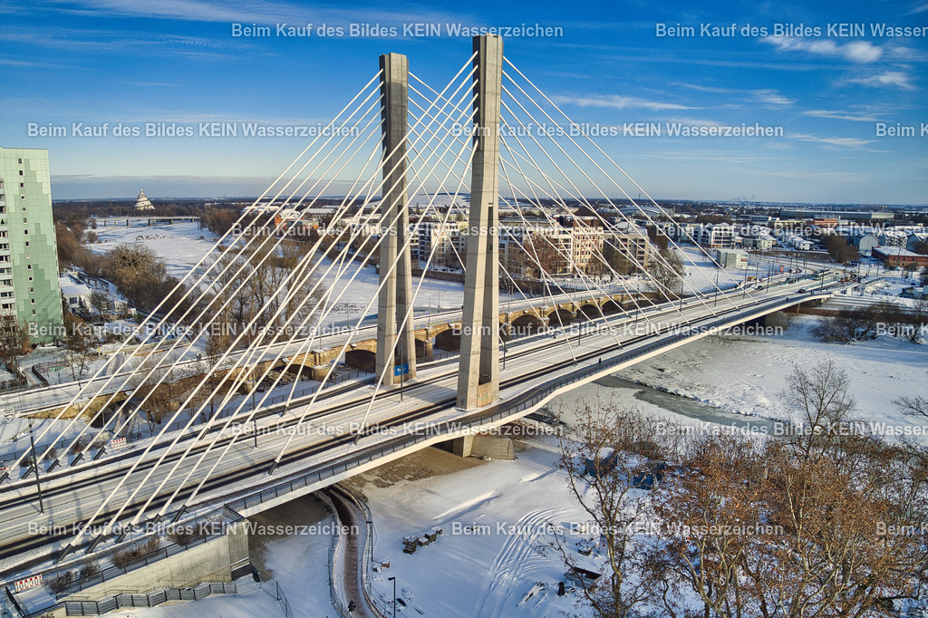 magdeburg_kaiser-otto-brücke_0007 | Das Luftbild zeigt die Kaiser-Otto-Brücke in Magdeburg aus geringer Höhe an einem winterlichen Tag. Die schneebedeckte Schrägseilbrücke hebt sich eindrucksvoll von der winterlichen Elblandschaft ab und macht die filigrane Konstruktion der Pylone und Seile besonders gut sichtbar. Aus der Drohnenperspektive werden die klaren Linien und die moderne Ingenieurarchitektur des Bauwerks eindrucksvoll hervorgehoben.Die Kaiser-Otto-Brücke wurde zwischen 2020 und 2024 als Teil des Ersatzneubaus des Strombrückenzuges errichtet. Das Bauwerk besitzt eine Gesamtlänge von 247,99 Metern und eine Breite von 27,10 Metern. Zwei 63 Meter hohe Pylone tragen den Brückenüberbau über 28 Schrägseile. Die Hauptöffnung überspannt die Alte Elbe mit einer Spannweite von 163,57 Metern. Die Brücke ist für Straßenverkehr, Straßenbahn, Radfahrer und Fußgänger ausgelegt und zählt zu den modernsten Brückenbauwerken Sachsen-Anhalts.Die winterliche Drohnenaufnahme verdeutlicht die technische Eleganz der Schrägseilkonstruktion und die präzise Ingenieurleistung des Bauwerks. Die schneebedeckte Landschaft verstärkt den Kontrast zur modernen Stahl- und Betonkonstruktion und macht die Kaiser-Otto-Brücke zu einem eindrucksvollen Beispiel zeitgemäßer Brückenbaukunst. - Realisiert mit Pictrs.com